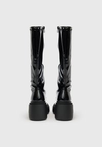 Crne čizme od lakirane kože do koljena s zaobljenim vrhom, teksturiranim bočnim šavovima i debelim crnim potplatom. Patentni zatvarač na vrhu.