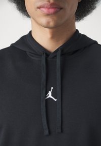 Jeune personne portant un sweat à capuche noir avec un logo Jumpman blanc et des cordons, visible du menton au haut de la poitrine sur fond blanc.