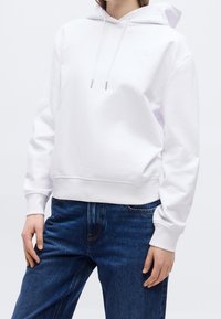 Personne portant un sweat à capuche blanc uni avec des cordons et un jean bleu, debout devant un fond neutre.