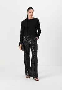 Haut noir à manches longues associé à un pantalon large en sequins noirs brillants. Le modèle tient une pochette noire et porte des sandales à talons. Texture lisse.