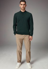 Camisa polo de manga larga verde oscuro con cuello y tres botones, combinada con pantalones beige y zapatillas blancas, de pie sobre un fondo neutro.