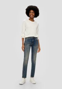 Biały sweter dzianinowy, niebieskie dżinsy z wyblakłym wzorem i białe trampki. Model stoi w swobodnej pozie na neutralnym tle.