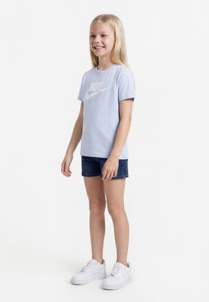 Ragazza sorridente in piedi, che indossa una t-shirt Nike azzurro chiaro, shorts di denim scuro e sneakers Nike bianche, su sfondo bianco uniforme.