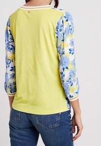 Vrouw in een gele blouse met blauw bloemenpatroon en 3/4 mouwen, gezien van achteren tegen een lichte achtergrond.