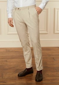 Beige Stoffhosen mit strukturiertem Finish, ausgestattet mit einem Knopfverschluss und zwei vorderen Falten. Getragen mit dunklen Slippern und burgunderfarbigen Socken.