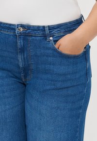 Tumši zili džinsi ar augstu jostas vietu, ar priekšējo kabatu, metāla pogām un smalkām šuvju detaļām uz šuvēm.