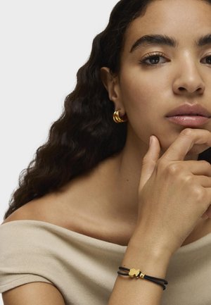 Pulsera negra con acentos dorados y forma única, llevada en una mano apoyada en el mentón. Blusa beige suave sin hombros y cabello oscuro ondulado.