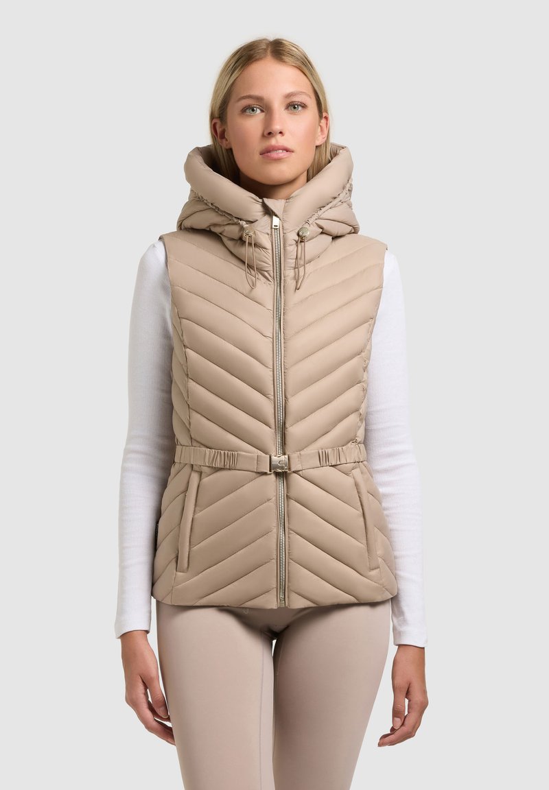 Blonde vrouw die een beige doorgestikte mouwloze jas met capuchon, riem en voorrits draagt over een witte top met lange mouwen tegen een effen achtergrond.