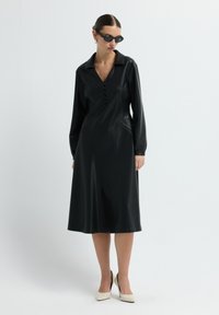 Žena v černých midi šatech s dlouhým rukávem a knoflíky na hrudi, s černými slunečními brýlemi a bílými vysokými podpatky, stojící na bílém pozadí.