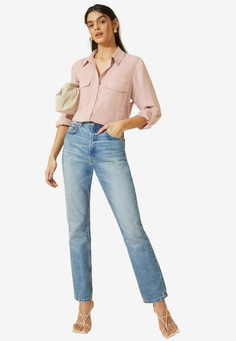 Helles rosa Hemd mit Knöpfen und zwei frontalen Taschen, kombiniert mit hoch taillierten blauen Jeans und beigen Sandalen mit Riemen. Mit einer strukturierten Handtasche.