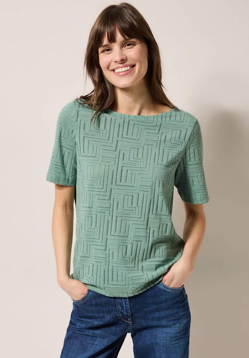 Haut mint vert à manches courtes avec un motif géométrique texturé, col rond et coupe décontractée, associé à un jean en denim bleu.