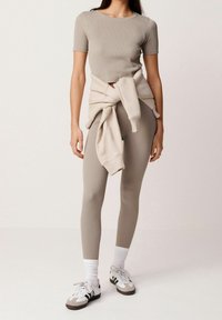 Jasnoszary prążkowany t-shirt, beżowe legginsy z wysokim stanem i białe sportowe trampki. Sweter zawiązany w talii dodaje faktury.