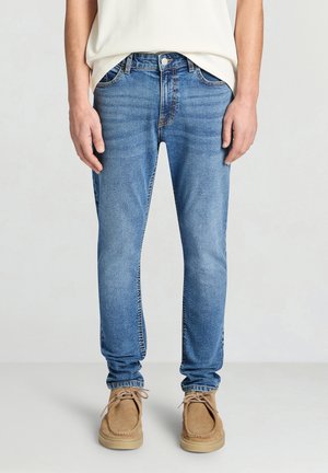 Jean en denim bleu clair à coupe slim, design à cinq poches, coutures visibles et fermeture par bouton en métal ; associé à des chaussures en daim couleur beige.