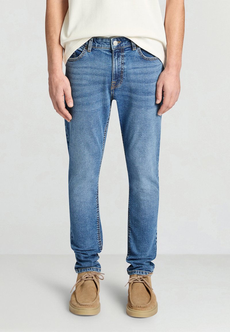 Jeans in denim azzurro chiaro con vestibilità slim, design a cinque tasche, cuciture visibili e chiusura con bottone metallico; abbinati a scarpe in suede color cammello.