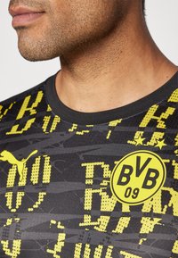 Camiseta atlética negra y amarilla con un patrón texturizado y el logotipo de Puma, con un cuello redondo y un emblema circular del club.