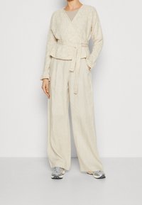 Beige wrap sweater with textured fabric and tie waist, paired with loose-fitting beige trousers. Sneakers feature grey and black accents.