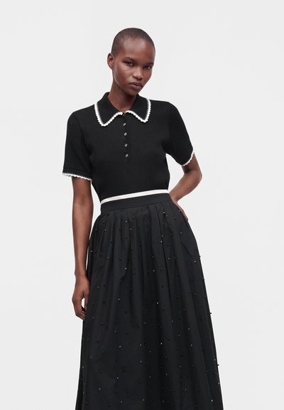 Femme aux cheveux courts portant un haut noir à manches courtes avec col et bordure blanche et une longue jupe noire avec de petits détails en perles.