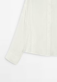 Chemise blanche à manches longues boutonnée avec des détails en dentelle sur le côté et un poignet structuré sur la manche.