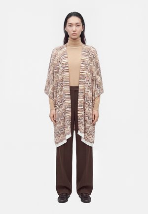 Un mannequin se tient devant un fond blanc uni, vêtu d’un pull à col roulé beige, d’un cardigan ouvert à motifs, d’un pantalon marron à jambes larges et de chaussures foncées.