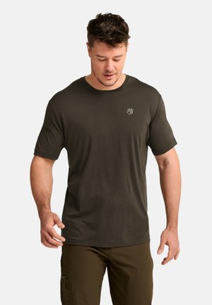 Uomo con camicia verde oliva scuro a maniche corte e pantaloni abbinati, che guarda verso il basso su uno sfondo chiaro e uniforme.