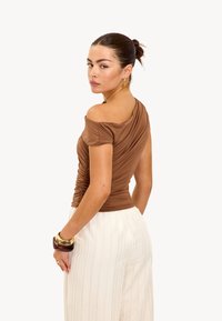 Bruine off-shoulder top met rimpeling, gecombineerd met witte geplooide broek. Een gouden armband om de pols voegt een contrasterende acccent toe.