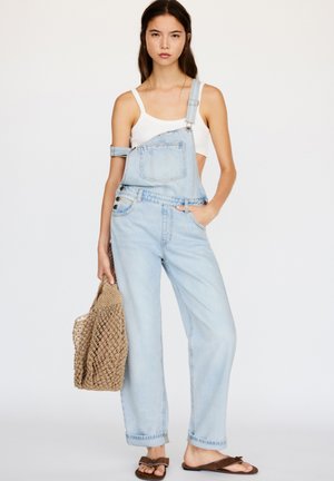 Lichtblauwe denim overall met een voorzak, verstelbare schouderbanden en vijf zakken, gecombineerd met een witte tanktop en bruine sandalen.