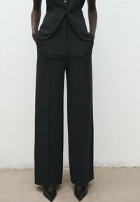 Pantalon large noir en tissu texturé, avec des poches latérales et une coupe ajustée, associé à des chaussures noires à bout pointu.