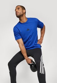 Blauwe sportt-shirt met een ronde hals en zwart Adidas logo. Het model draagt zwarte broeken met witte strepen en houdt een kettlebell vast.