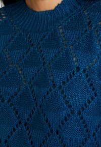 Sweter dzianinowy w ciemnoniebieskim kolorze z teksturowanym wzorem w romby i detalami w otwartym splocie. Posiada ściągacz przy okrągłym dekolcie.