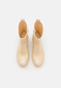 Beige enkellaarsjes voor de regen, gemaakt van glanzend rubber, met een gladde textuur, een ronde neus en twee trekflappen met zilveren hardware.
