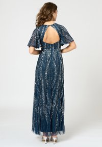 Laivansinis floor-length mekko, jossa on läpikuultavat, liehuvat hihat ja avonainen selkä. Koristeltu hopeakuosisilla yksityiskohdilla, joissa on teksturoitu, virtaava muotoilu.