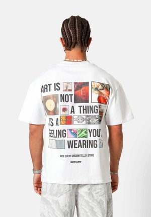 Hombre con cabello trenzado que lleva una camiseta blanca con imágenes coloridas y texto que dice "EL ARTE NO ES UNA COSA, ES UN SENTIMIENTO QUE LLEVAS PUESTO."