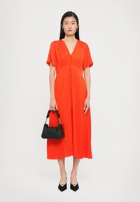 Red midi dress with short sleeves, V-neck, and button details down the front. Features a textured fabric. Paired with a black handbag.