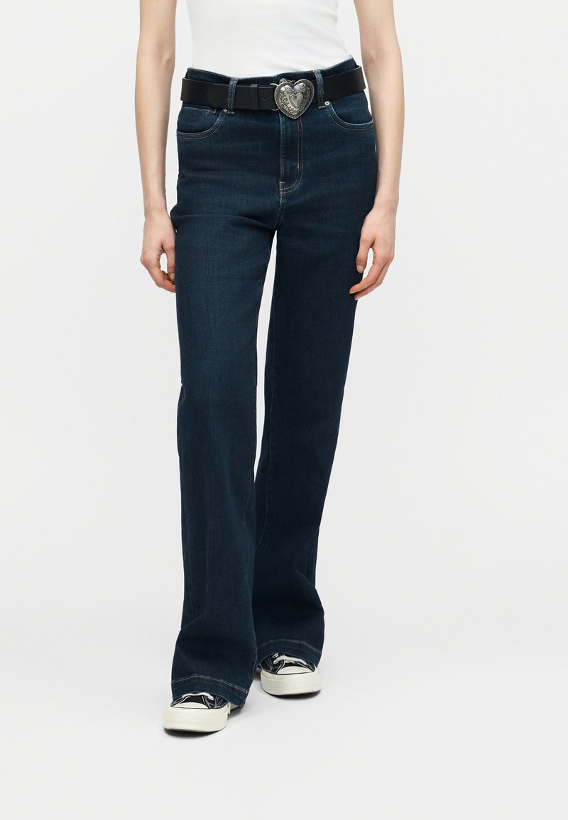 Άτομο που φοράει σκούρο μπλε τζιν καμπάνα, λευκό τοπ, μαύρη ζώνη με ασημένια πόρπη σε σχήμα καρδιάς και ασπρόμαυρα παπούτσια.