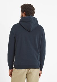 Man with curly hair wearing a dark navy hoodie and beige pants, facing away against a plain white background.