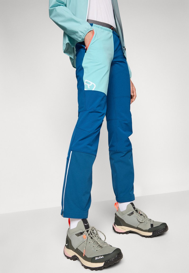 Blaue und türkisfarbene Wanderhosen mit schlanker Passform, ausgestattet mit Taschen und einem weißen Logo. Kombiniert mit grauen und schwarzen Trailschuhen mit Akzenten.