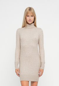 Mujer rubia vistiendo un vestido de punto beige con cuello alto y detalles de botones en los puños de las mangas, de pie frente a un fondo blanco.