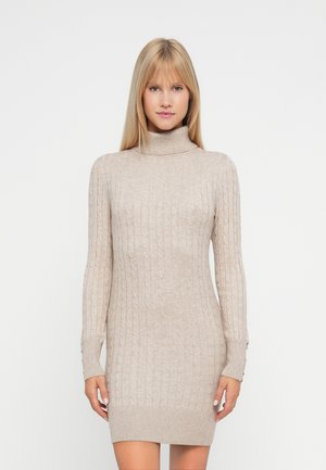 Mujer rubia vistiendo un vestido de punto beige con cuello alto y detalles de botones en los puños de las mangas, de pie frente a un fondo blanco.