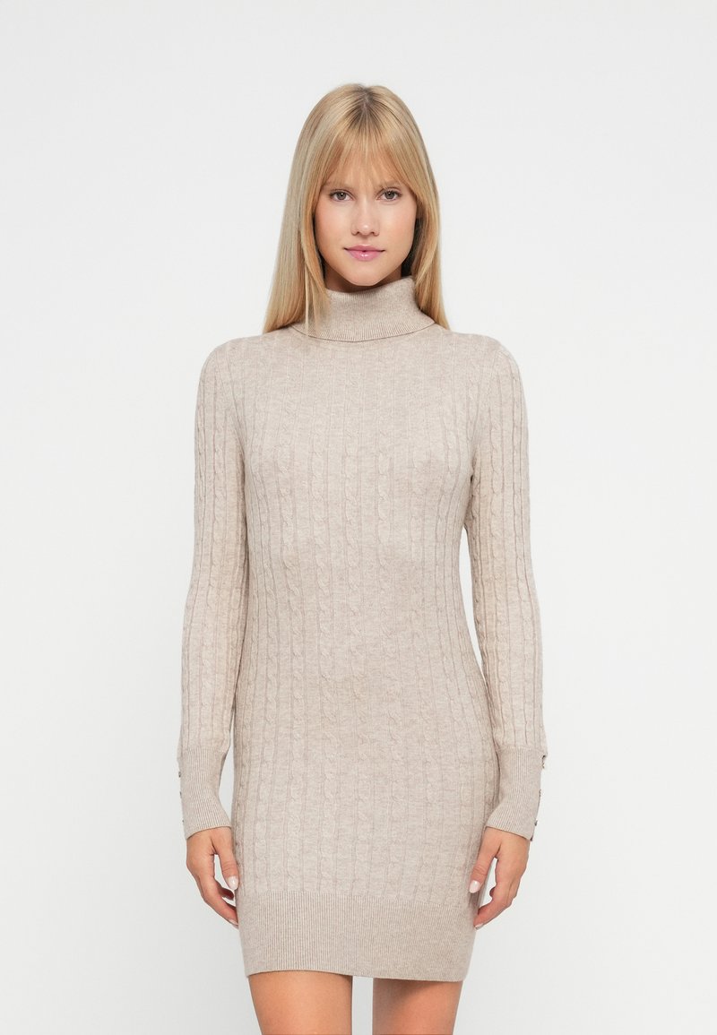 Mujer rubia vistiendo un vestido de punto beige con cuello alto y detalles de botones en los puños de las mangas, de pie frente a un fondo blanco.