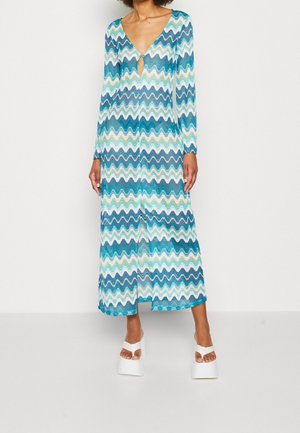Vrouw draagt een lange jurk met lange mouwen tot aan de enkel met blauwe, witte en groene zigzagpatronen en witte platformsandalen.
