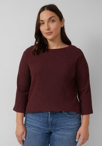 Burgunderfarbener strukturierter Strickpullover mit weitem Ausschnitt und Dreiviertelärmeln, kombiniert mit blauen Jeans. Sichtbare Seitennähte.