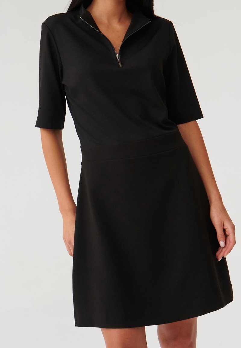 Femme portant une robe noire mi-longue avec des manches jusqu'aux coudes et un col zippé à l'avant, sur un fond uni.
