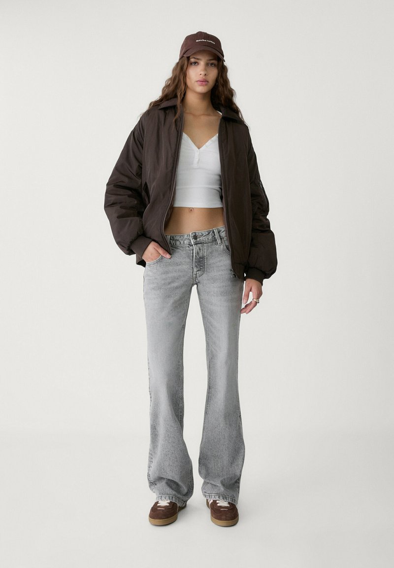 Veste bomber marron, crop top blanc, jean évasé gris et baskets marron. Les accessoires comprennent une casquette marron avec des détails textuels.