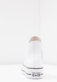 High-top white sneaker made of smooth synthetic material. Features a black-striped sole and a logo patch on the outer heel.