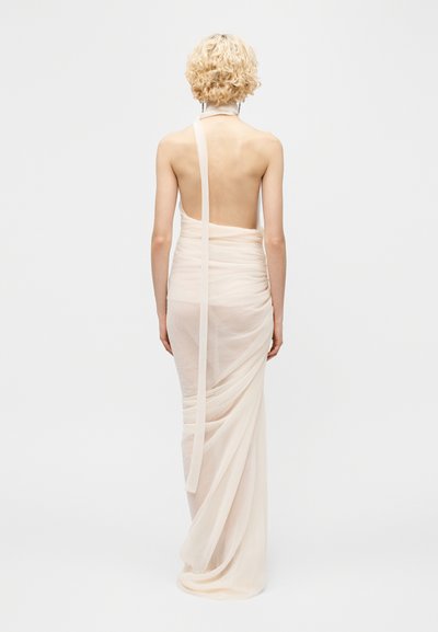 Person with short curly blonde hair wearing a floor-length, backless, sheer pale pink gown standing against a plain white background.