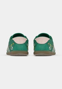 Πράσινα αθλητικά παπούτσια με λευκές λεπτομέρειες, διακοσμημένα με λουλουδένια μοτίβα και υφή που θυμίζει σουέντ. Η πίσω όψη δείχνει χαμηλό προφίλ και κυρτή φτέρνα.