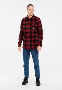 Rood en zwart geruit flanellen shirt met een knoopsluiting, voorzien van een borstzak en een gladde textuur, gecombineerd met blauwe jeans en zwarte schoenen.
