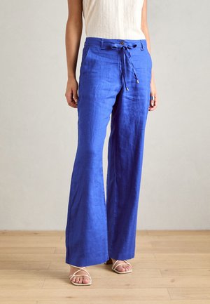 Blauwe linnen broek met wijde pijpen en een taille met koord, gedragen met een wit mouwloos topje en witte sandalen met bandjes.