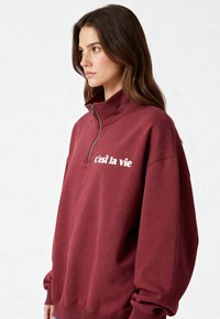 Sweatshirt bordeaux à fermeture éclair avec col montant, comportant le texte blanc "c'est la vie" sur le devant. Mélange doux de coton, coupe décontractée, poignets côtelés.