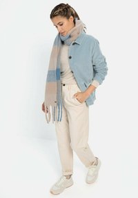 Lichtblauwe corduroy jas met knopen, beige gestructureerde sjaal met blauwe strepen, crèmekleurig topje, lichtgekleurde broek en grijze sneakers.